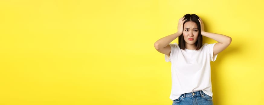 Asian woman holding hands on head and looking sad, having a problem, standing anxious against yellow background