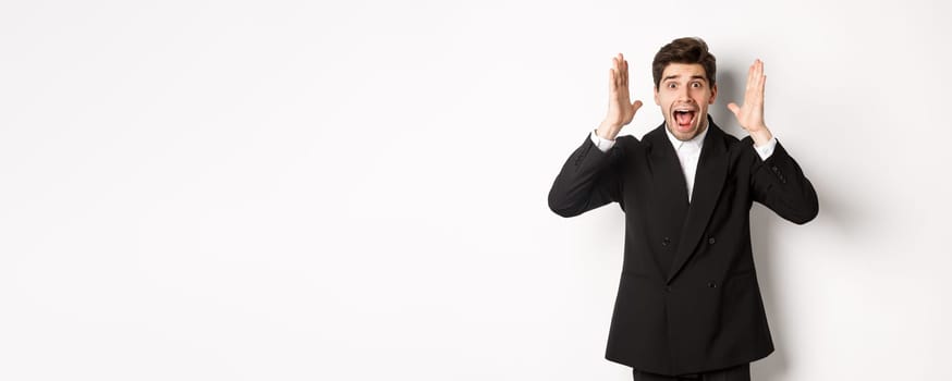 Frustrated and concerned man in black suit, screaming in panic and looking at something shocking, standing over white background