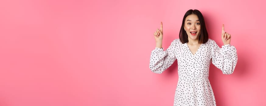 Amazed brunette asian girl pointing fingers up, showing advertisement, demonstrate spring promo offer, standing over pink background