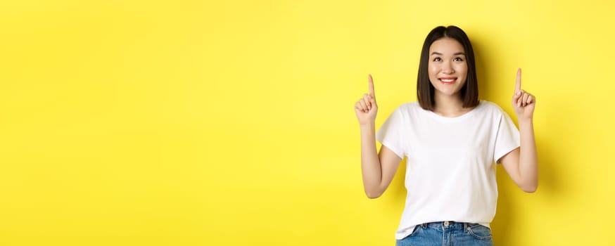 Beauty and fashion concept. Beautiful asian woman in white t-shirt pointing fingers up, standing over yellow background
