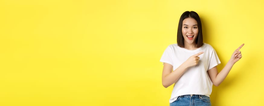 Beauty and fashion concept. Beautiful asian woman in white t-shirt pointing fingers right, demonstrate logo standing over yellow background.