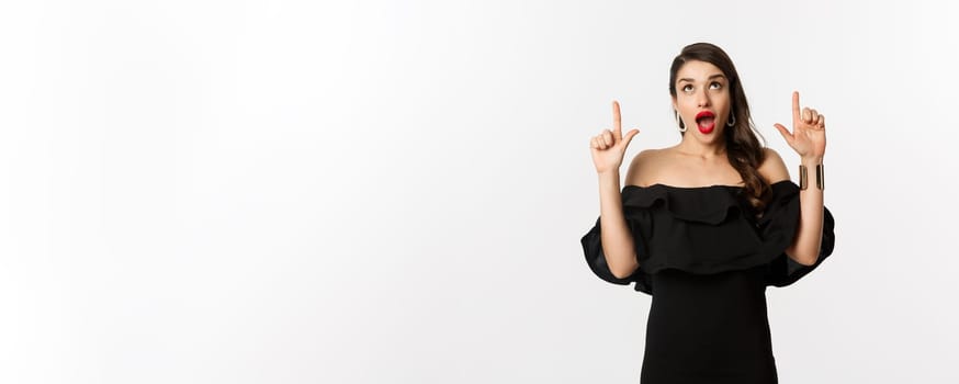 Fashion and beauty. Amused woman in black dress looking and pointing fingers up at promo offer, standing over white background