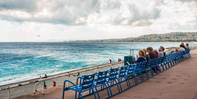 People are relaxing on blue bench on the beach of Mediterranean sea, Nice, France.