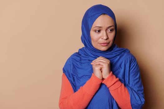 Middle-aged Muslim woman in blue hijab and hopeful look, with hands folded on chest level, prays Allah for peace. Islam