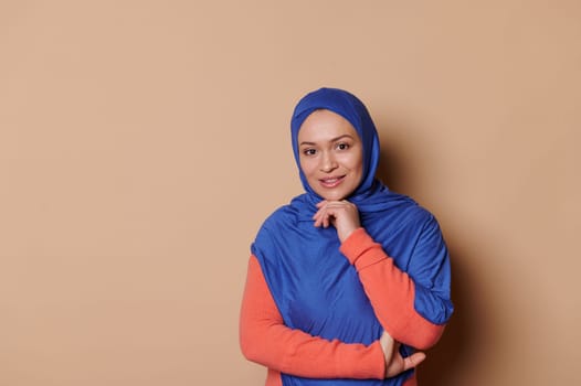 Confident elegant charming middle-aged Arab Muslim woman in blue hijab, looking at camera, isolated on beige background