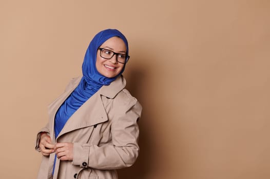 Stylish elegant confident Muslim woman in blue hijab and eyeglasses, smiling looking aside on isolated beige background