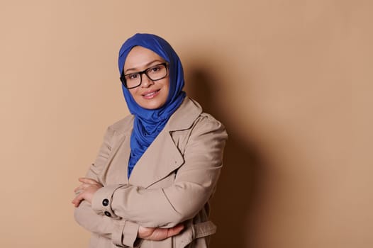 Smiling Muslim woman in elegant strict outfit, blue hijab and stylish spectacles, posing arms crossed, beige background