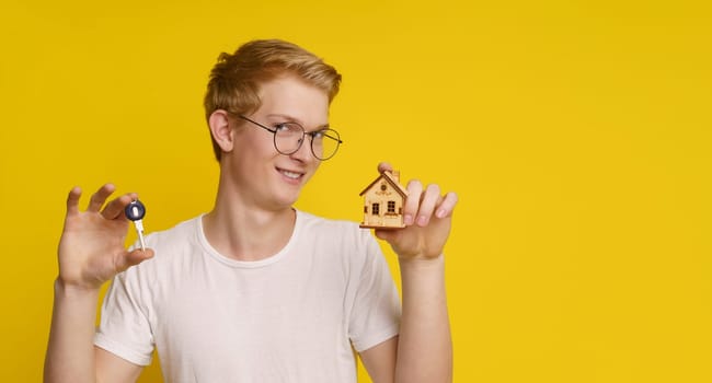 Young man holding a home architectural model, maquette and key against yellow background, showcasing the achievement of home ownership and success in the real estate market.
