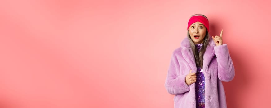 Fashion and shopping concept. Smiling elderly woman having an idea. Stylish old lady in purple fake fur coat raising index finger, suggesting plan, standing excited on pink background