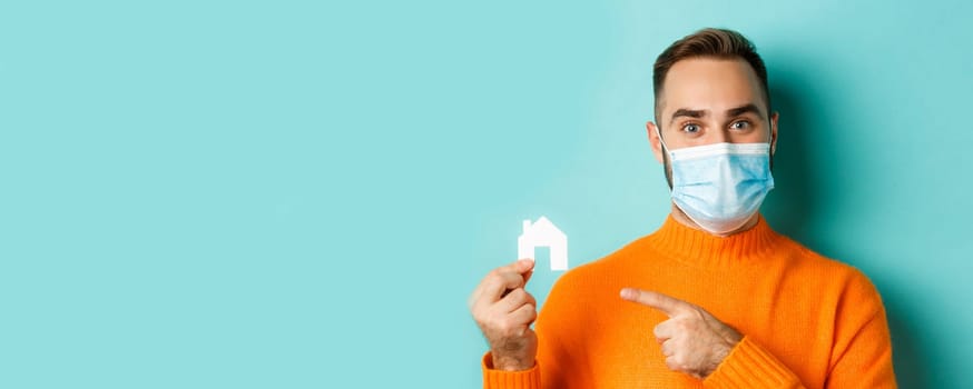 Real estate and coronavirus pandemic concept. Close-up of adult man in medical mask pointing at small paper house, standing over light blue background