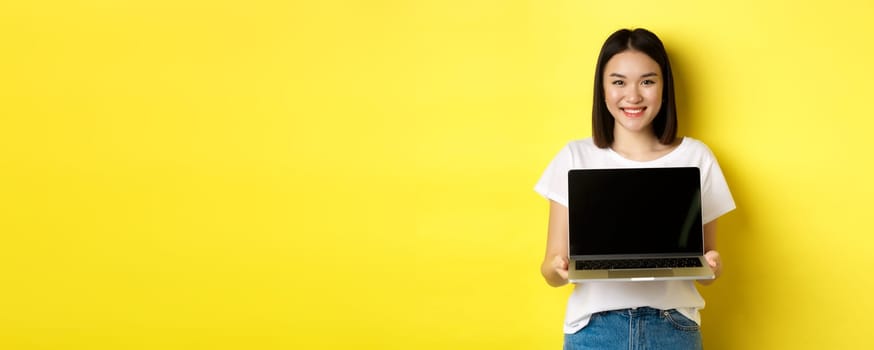 Young asian woman demonstrate online offer, showing laptop screen and smiling, standing over yellow background
