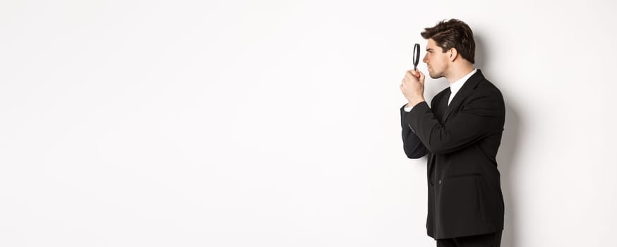 Profile shot of handsome businessman in black suit, looking through magnifying glass and searching for something, standing over white background