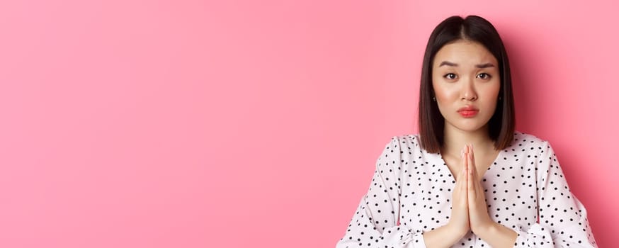 Beauty and lifestyle concept. Sad asian woman asking for help, begging with hands in plead gesture, staring at camera, need favour, standing over pink background.