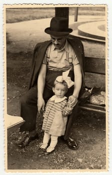 A vintage photo shows small toddler girl and father. Toddler shows sad face.