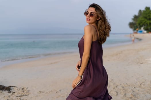 Stylish romantic tender sensual woman in silk dress and sunglasses on the beach at sunset