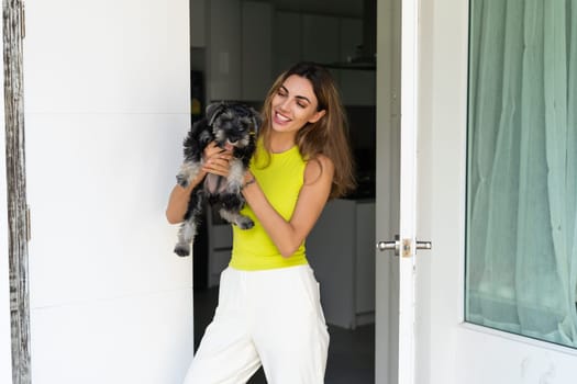 Welcome. Portrait of cheerful woman standing in doorway of modern apartment, greeting visitor and inviting guest to enter her home, holding a puppy