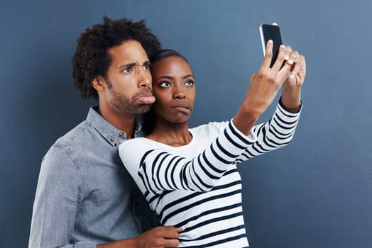 Being weird together is our thing. a young couple making faces while taking a photo of themselves with a cellphone on a gray background.