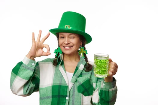 Cheerful woman in green party hat, holds a mug of Irish beer, winks, shows Ok hand sign at camera. Saint Patrick's Day