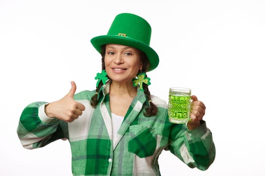 Stylish woman wearing clover leaf earrings and party hat, holds a mug of Irish green beer and shows thumb up at camera