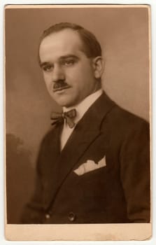 Vintage studio portrait shows man wears jacket and bow tie. Antique black white photo with sepia tint.