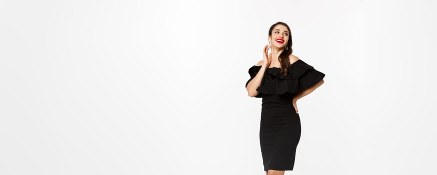 Celebration and christmas holidays concept. Beautiful woman in black dress with gifts and looking surprised, standing over white background.