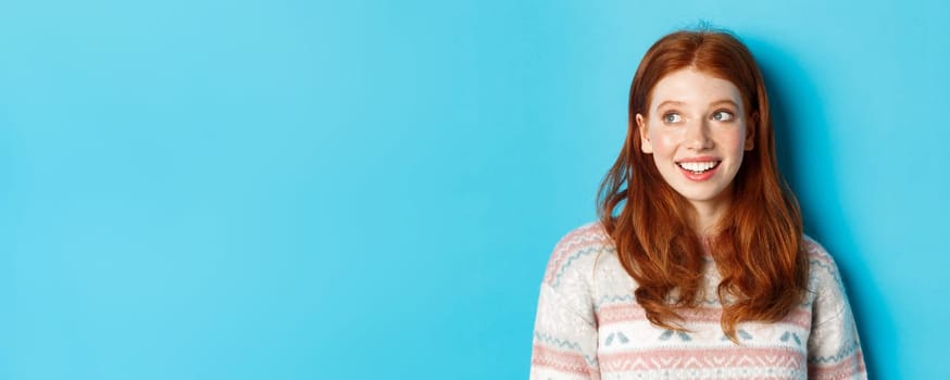 Close-up of dreamy redhead girl in winter sweater looking left, smiling and staring at promo offer, standing against blue background