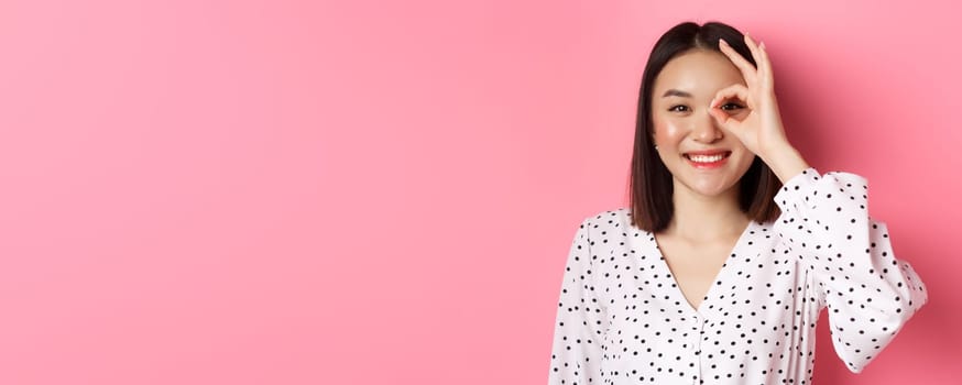 Beauty and lifestyle concept. Close-up of cute smiling asian girl showing okay sign on eye, standing over pink background in dress.
