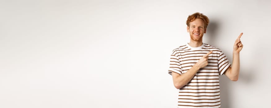 Handsome redhead man showing logo, pointing fingers right and smiling, standing over white background