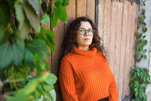 Autumn portrait of an attractive young woman in stylish glasses in a knitted fashionable orange sweater on background of leaves and big door . Girl walks in city - copy space and empty space for text