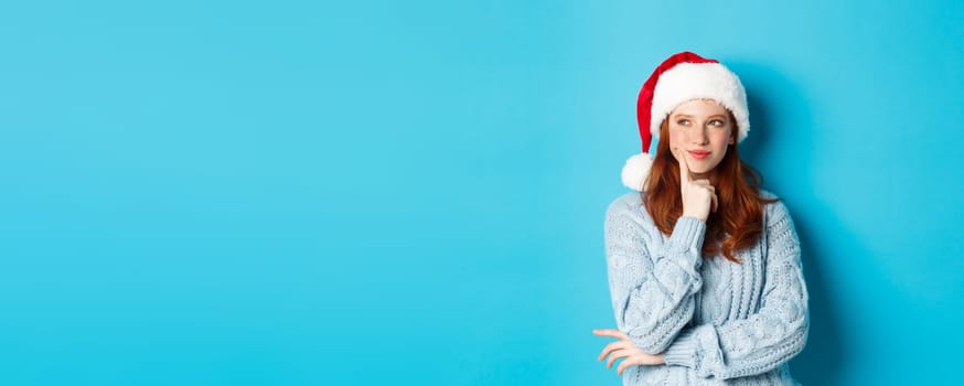 Winter holidays and Christmas Eve concept. Silly redhead girl with freckles, wearing santa hat and thinking, planning New Year celebration, standing over blue background