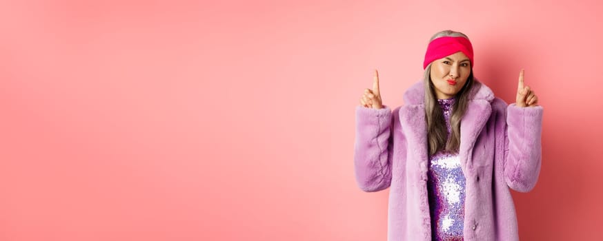 Disappointed stylish asian lady pointing fingers up. Senior woman in trendy fake fur coat grimacing skeptical and pointing fingers up, dislike promo, standing over pink background