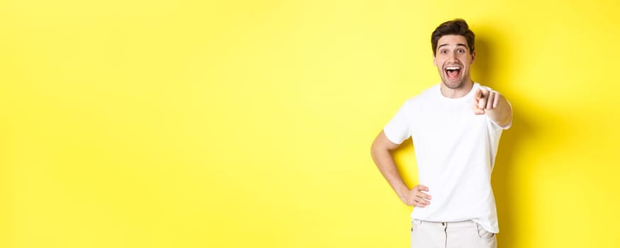 Happy guy pointing finger at camera and laughing, check out something, standing over yellow background