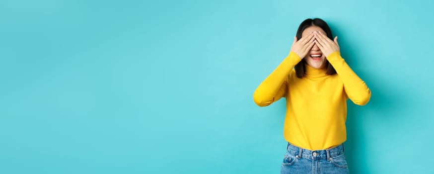Cheerful asian girl in yellow pullover waiting for surprise, playing hide n seek and smiling, expecting gift with eyes closed, standing over blue background