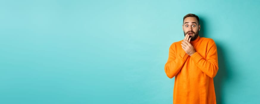 Scared and shocked guy looking at camera, standing in orange sweater against turquoise background