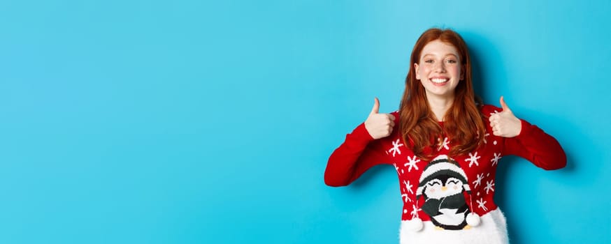 Winter holidays and Christmas Eve concept. Happy smiling girl with red hair, showing thumbs up in approval, praise good product, recommending, standing in xmas sweater