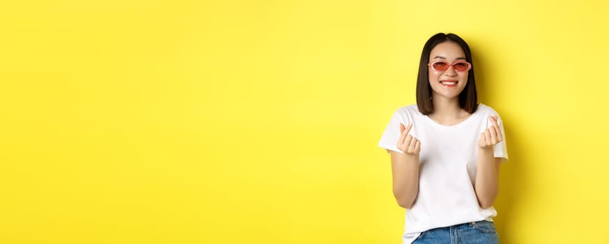 Fashion and lifestyle concept. Attractive asian woman in stylish sunglasses, showing finger hearts and smiling happy at camera, standing over yellow background