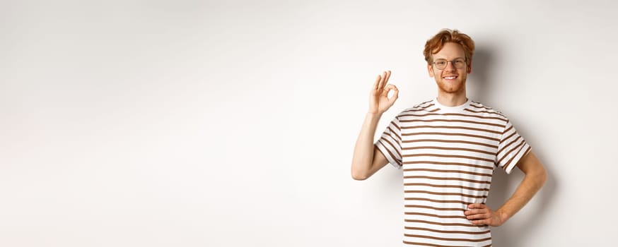 Confident smiling man with red hair assuring you, showing OK sign, guarantee quality, recommending something good, standing over white background