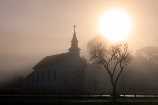 Morning sun through dense fog over small historic church and bare tree