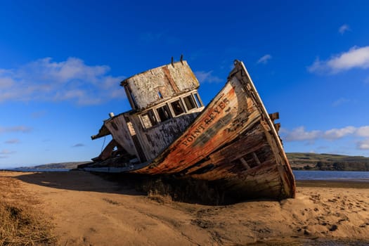 Historic wooden fishing boat wreck on sandy beach in sun and blue sky