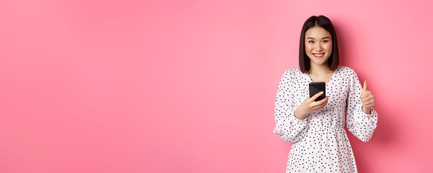 Online shopping and beauty concept. Satisfied asian female customer showing thumbs-up, making purchase in internet on smartphone, standing over pink background.