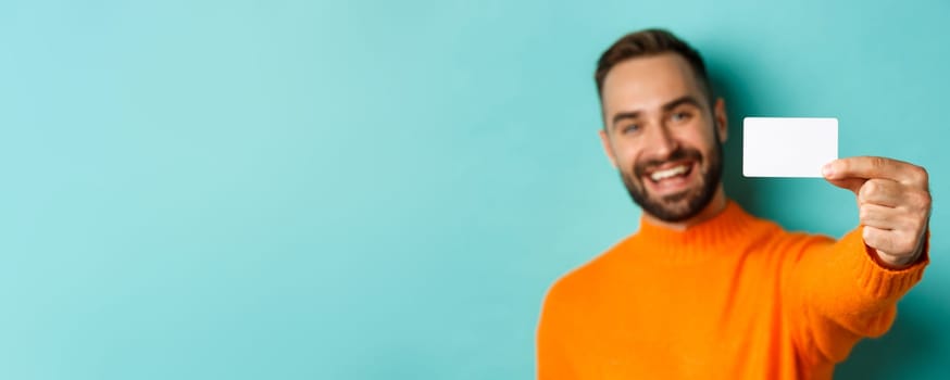 Close-up of handsome caucasian man going on shopping, showing credit card and smiling, standing over turquoise background