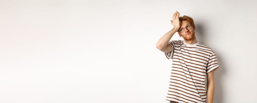 Tired young man with red hair and glasses, roll eyes and making facepalm bothered, standing over white background