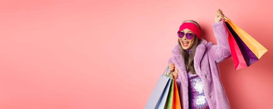 Funky and cool asian senior woman in fashionable clothes dancing while going shopping on sales, holding shop paper bags and having fun, pink background