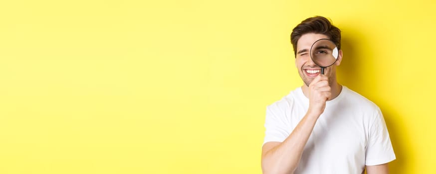 Close-up of young man looking through magnifying glass and smiling, searching something, standing over yellow background