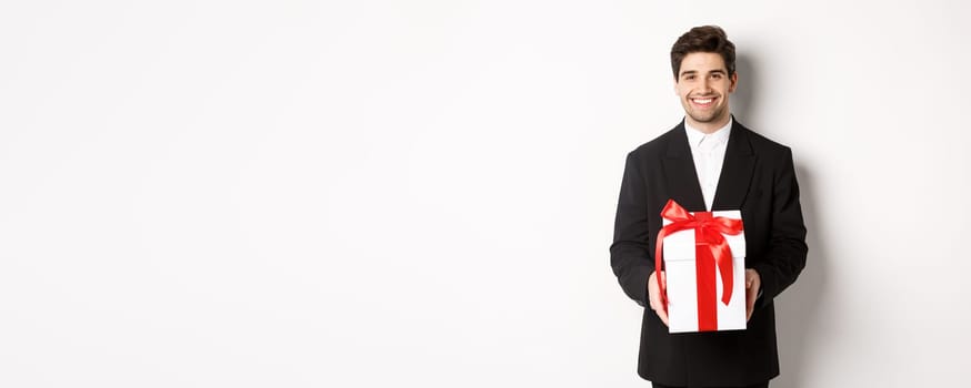 Concept of christmas holidays, celebration and lifestyle. Handsome man in black suit, have romantic present, holding gift in a box and smiling, standing against white background