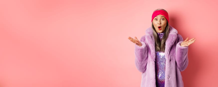 Fashion and shopping concept. Surprised elderly asian woman in stylish fake-fur coat and headband looking amazed at camera, rejoicing of good news, pink background
