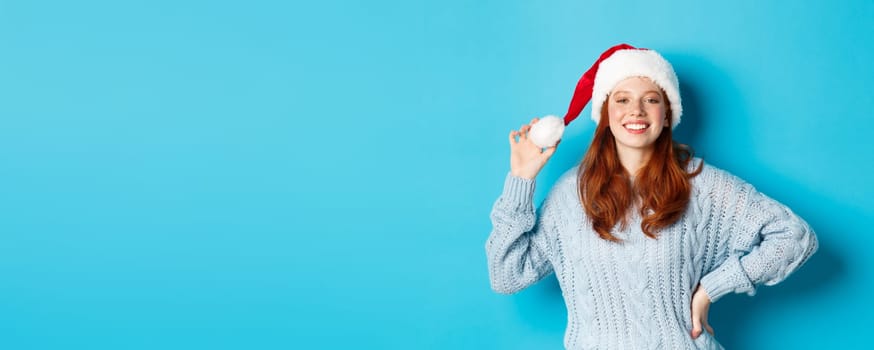 Winter holidays and Christmas Eve concept. Cheerful redhead teenage girl wearing Santa hat and smiling satisfied, standing in sweater against blue background