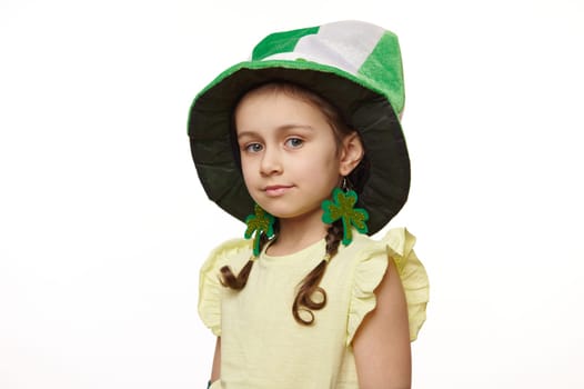 Stylish little Irish baby girl wearing clover leaves earrings and green hat, smiles looking at camera, white background
