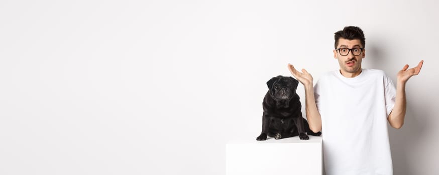 Image of confused man in glasses raising hands and shrugging complicated, standing near black pug dog over white background