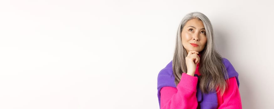 Close up of thoughtful asian female with grey hair, looking up and thinking, making decision, standing over white background